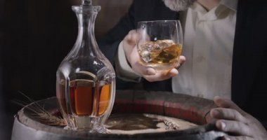 Takım elbiseli ve sakallı bir adam (şarap garsonu) eski bir viski fıçısından buzlu bir bardak viski alır ve viski kokusunu yaymak için bardağı sallar. Yavaş çekim 4K, 150 fps, Blackmagic Ursa Pro G2. 