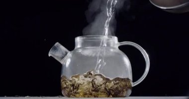Tam bir çay demleme işlemi. Kaynar su, büyük yaprak çayı, koyu kahverengi arka plan, yavaş çekim 150 fp içeren cam bir çaydanlığa dökülür.