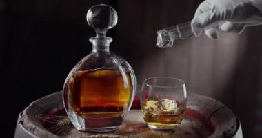 Bir buz küpü yavaşça viski bardağının üstüne dökülüyor. Eski bir viski fıçısının üstüne. İçkili bir sürahi yanında duruyor, koyu kahverengi bir arka plan. Blackmagic Ursa Pro G2
