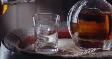 Bir buz küpü kadehe yavaşça düşer, eski bir viski fıçısının üzerinde durur, şişeden gelen viski hemen bardağa dökülür. İçkili bir sürahi yanında duruyor, koyu kahverengi bir arka plan. Blackmagic Ursa Pro G2.