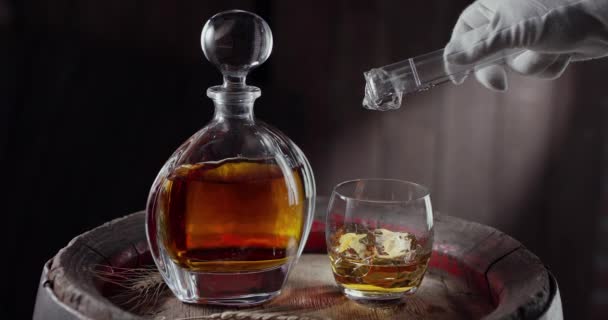 アイスキューブは ヴィンテージのウイスキー樽の上にゆっくりとウイスキーのグラスに落ちます 飲み物のあるデカンタは近くに立っており 暗褐色の背景をしている Blackmagic Ursa Pro — ストック動画
