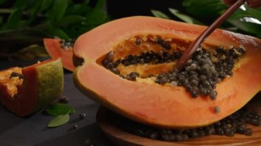 Yarım papaya meyvesi ve tropikal bitki yapraklarıyla birlikte antika bir masa üzerinde tohum. Tahta kaşıkla bir el tohumlardan papaya meyvesini temizler. Kameranın soldan sağa yatay yavaş hareketi. 