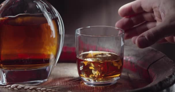 男人的手拿着一杯威士忌和冰块 站在一个老式的威士忌桶上 旁边站着一个酒壶 背景是深褐色的 — 图库视频影像