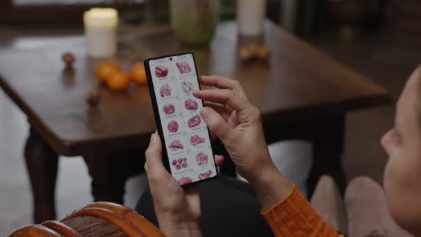 自宅でスマートフォンを使って料理を注文する スマートフォンのアプリを使ってオンラインストアで肉や魚を選ぶ女性 家の夜の家具 — ストック動画