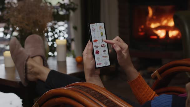 自宅でスマートフォンを使って料理を注文する 女性はスマートフォンのアプリを使ってオンラインストアで市場食品を選択します 暖炉の火で家の夜の家具 — ストック動画