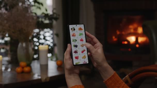 在家里用智能手机点菜 一个女人用智能手机上的应用程序在网上商店里挑选蔬菜 炉火熊熊燃烧的家居装饰 — 图库视频影像