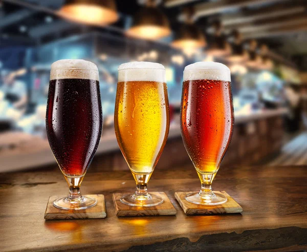 三杯不同的冰镇啤酒的冰镇玻璃杯放在木制桌子上 后面是模糊的酒吧内部 — 图库照片