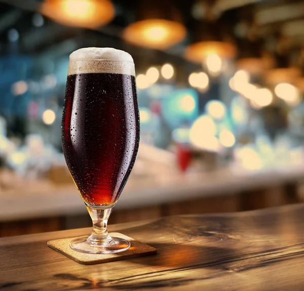 木制桌子上有一杯冰镇的黑啤酒 后面是模糊的酒吧内部 — 图库照片