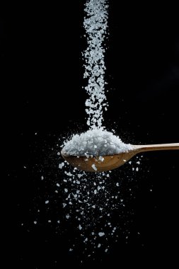 Siyah arka planda yenilebilir tuz kristalleri tahta kaşığa dökülüyor..