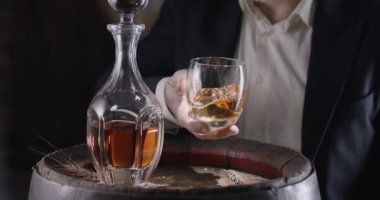 Man in a suit and beard (sommelier) takes a glass of whiskey on ice from a vintage whiskey barrel and shakes the glass to spread the smell of whiskey. Slow motion 4K