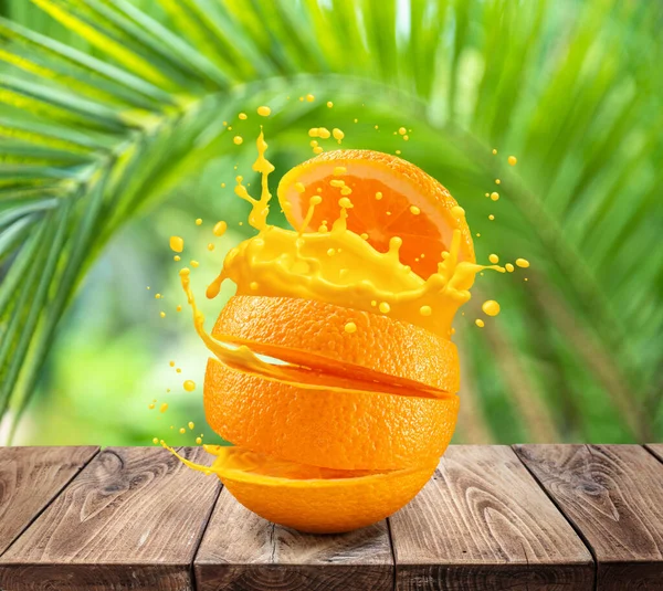 Sliced orange fruit splashing about orange juice on the wooden table. Green palm leaves at the background.