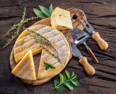 Head of  sheep milk cheese with fresh herbs and cheese knives on wooden background. 