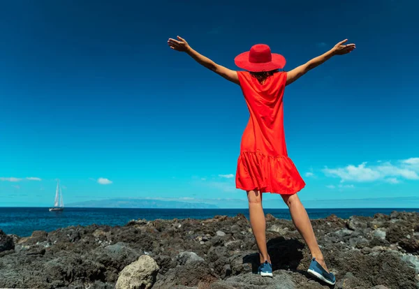 穿着红色衣服的快乐女人张开手站在岩石海岸上 平静的海水和蓝天背景 — 图库照片