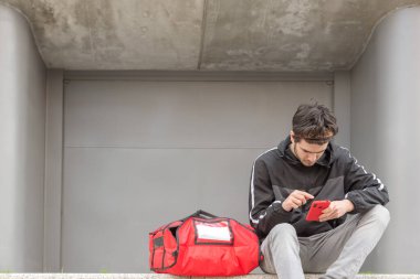 Kırmızı çantalı teslimatçı, bir sonraki siparişi vermek için cep telefonunu kullanıyor.