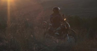 Miğferli adam Enduro motorsikletinde oturuyor ve vadide gün batımını izliyor.