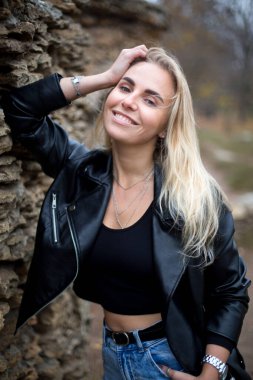 Siyah deri ceketli güzel beyaz kadın sonbahar parkında taş duvarın yanında duruyor.