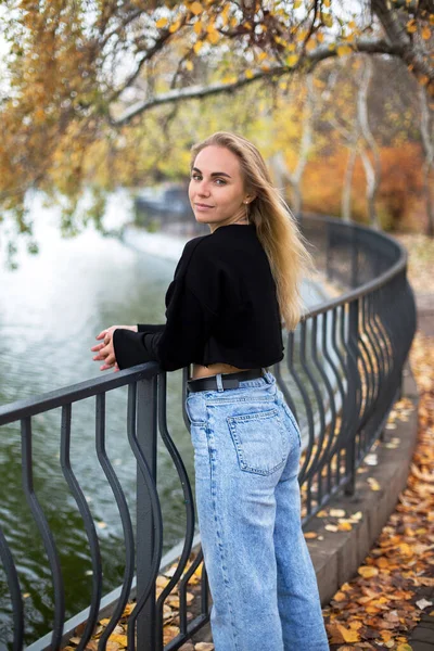 Güz parkında göletin kenarında yürüyen siyah kazaklı güzel beyaz kadın.