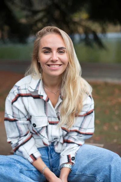 Beyaz flanel gömlekli güzel beyaz sarışın bir kadın sonbahar parkındaki dalda oturuyor.