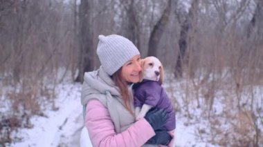 Kadın, karlı kış ormanında ceketli köpek yavrusuyla sarılıyor.