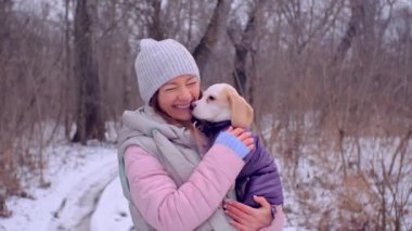 Kadın, karlı kış ormanında ceketli köpek yavrusuyla sarılıyor.