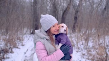 Kadın, karlı kış ormanında ceketli köpek yavrusuyla sarılıyor.