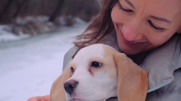 冷凍川の近くの雪中銀行に可愛いビーグル子犬とジャケットの抱擁の女性 — ストック動画