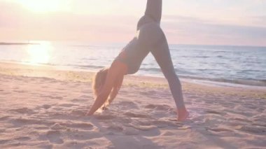 Orta yaşlı beyaz kadın, taytlı, denizin önündeki kumsalda yoga egzersizi yapıyor.