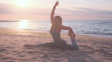 Orta yaşlı beyaz kadın, taytlı, denizin önündeki kumsalda yoga egzersizi yapıyor.