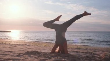 Orta yaşlı beyaz kadın, taytlı, denizin önündeki kumsalda yoga egzersizi yapıyor.