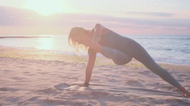Orta yaşlı beyaz kadın, taytlı, denizin önündeki kumsalda yoga egzersizi yapıyor.