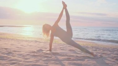Orta yaşlı beyaz kadın, taytlı, denizin önündeki kumsalda yoga egzersizi yapıyor.