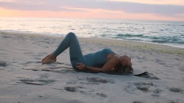 Orta yaşlı beyaz kadın, taytlı, denizin önündeki kumsalda yoga egzersizi yapıyor.