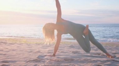 Orta yaşlı beyaz kadın, taytlı, denizin önündeki kumsalda yoga egzersizi yapıyor.