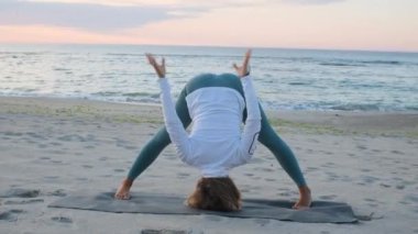 Orta yaşlı beyaz kadın, taytlı, denizin önündeki kumsalda yoga egzersizi yapıyor.