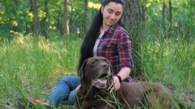 Kırmızı ekose gömlekli beyaz bir kadın yeşil parkta yaşlı bir labrador köpeğiyle oynuyor.