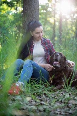 Kırmızı ekose gömlekli beyaz bir kadın yeşil parkta yaşlı bir labrador köpeğiyle oynuyor.