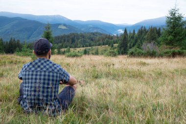 Yürüyüş yapan sakallı adam çimenlerde oturuyor ve dağ manzarasına bakıyor. Şapka, şort ve ekose gömlek giymişti.