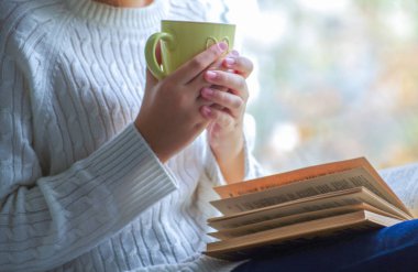 Kız elinde bir fincan çay tutuyor ve pencerenin yanında kitap okuyor.
