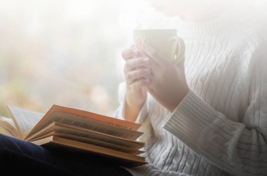 Elinde bir fincan çay tutan ve pencerenin yanında kitap okuyan bir kadın.