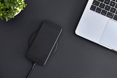 Yuvarlak indüksiyon kablosuz şarj cihazı kullanarak akıllı telefon bataryasını şarj ediyorum. Ofis işyerini tepeden tırnağa görmek. Modern teknoloji, kablosuz aygıt ve enerji aktarımı.