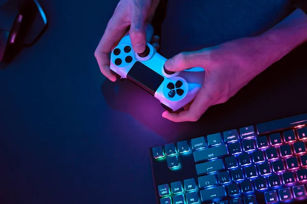 Bilgisayarı kontrol etmek için kontrolör kullanan erkek eller. Gökkuşağı renkleri arka plandaki bilgisayar klavyesini aydınlattı. Profesyonel bilgisayar oyunu, esport işi ve online dünya konsepti.