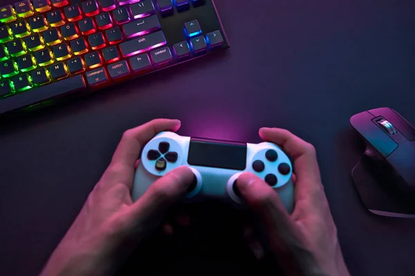 High angle view of male hand using game controller above dark desk with computer input devices. Professional computer game playing, esport business and online world concept.