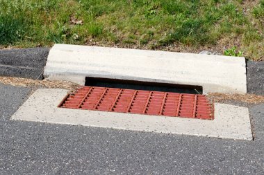 Banliyö sokağının yüzeyi kırmızıya boyanmış metal fırtınası su ızgarası açık havada, arkada çimen var. Banliyöde ve sokakta çimentoyla çevrili kırmızı boyanmış metal kanalizasyon ızgarasına yakın plan.