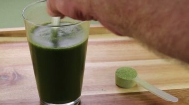 Daha önce karıştırılmış organik kereviz tozunu kadehte aloe vera suyu ile karıştıran bir erkek el. Kuru organik kereviz tozu kepçesini kereviz tozuyla karıştırılmış bir bardak aloenin yanında ölçüyor..