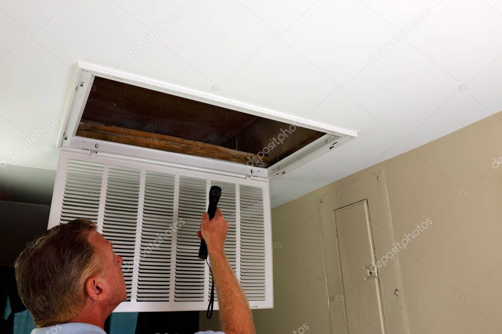 Flashlight shined into an air intake vent of a home HVAC system by an adult white male in an entryway. Adult male looking into an air intake vent with open grid after removing home HVAC air filter