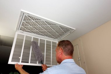 Mature male dusting with a gray dust wand a white grid of a home furnace air intake vent. Interior white grid of an HVAC air intake vent being dusted with a dust wand by a mature caucasian male
