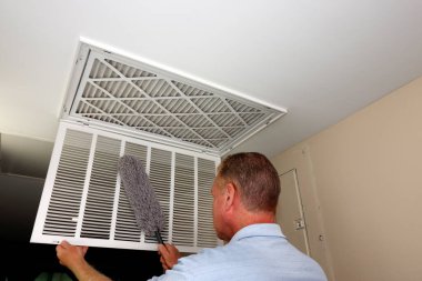 Mature male dusting with a gray dust wand a white grid of a home furnace air intake vent. Interior white grid of an HVAC air intake vent being dusted with a dust wand by a mature caucasian male clipart