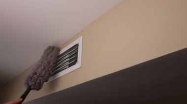 Human Hand Dusting a Small Air Duct Vent on a Home Wall. Small white rectangle furnace vent grill being dusted by a male hand with a long gray dusting wand. Wall air duct vent near a ceiling dusted with a long gray dust wand by an adult male hand