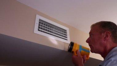 Man Examining Inside a Home Wall Air Duct Vent with a Flashlight. Adult white male with a flashlight inspecting the inside of an air duct in a home wall near a ceiling. Home air vent duct on a wall being inspected by a mature man with a flashlight.