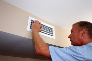 White rectangle air vent on a beige wall with a males hand on left side of the outflow furnace register. Mature male with left hand on and inspecting white home furnace air duct vent in a modern hom